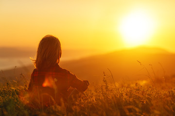 Woman sits with her back in the field and admires the sunset in the mountains.