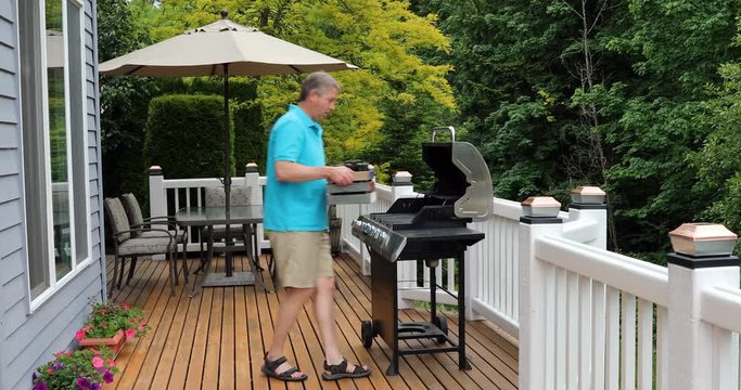 Mature man taking wooden box filled with bottled beer to BBQ cooker on outdoor cedar deck. 