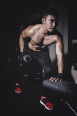 Fototapeta na wymiar Asian young man with dumbbells flexing muscles in gym, Dark lighting low key concept
