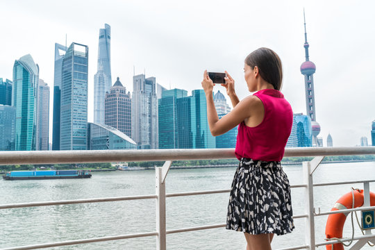 Asian business woman taking phone picture of Pudong skyline while on Shanghai ferry cruise towards the financial district center, China. Female tourist looking at view taking snapshots.