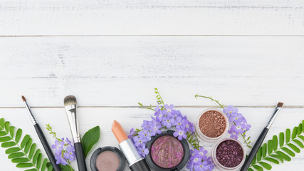 Purple flowers, green leaves, cosmetics and make up brushes on white wood background with copy space