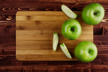 Green apples with slices and bamboo cutting board on brown plank, top view, copy space.