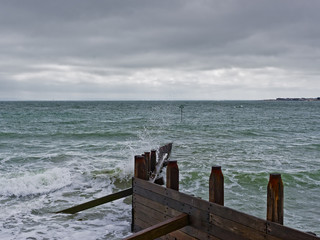 West Wittering beach on a windy day, under a grey sky, waves crash against a partially submerged timber groynes.