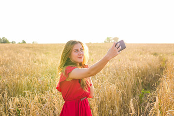 technology, summer holidays, vacation and people concept - smiling young woman in red dress taking selfie by smartphone on cereal field