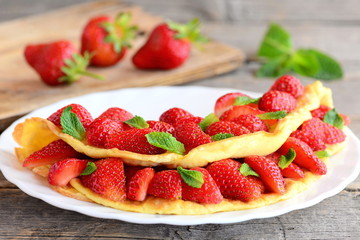 Breakfast souffle omelette with strawberries. Fried omelette filled with fresh strawberries slices and garnished with mint on a plate. Fresh strawberries on old wooden table. Closeup