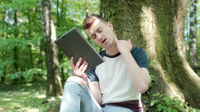 Young man being bitten by insects while sitting in the park and using tablet, steadycam shot
