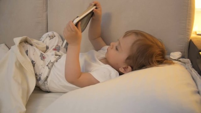 Toddler boy lying in bed and watching video on digital tablet