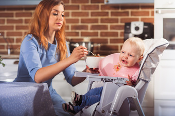 Little baby girl sits in high chair and won't  to eat the food and cry. Mother feeding her with spoon. Family and motherhood concept. Horizontal 