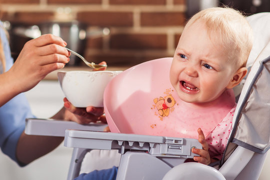 Little Baby Girl Sits In High Chair And Won't  To Eat The Food And Cry. Mother Feeding Her With Spoon. Family And Motherhood Concept. Close Up. Horizontal 