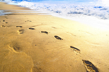 Footprints and a sea wave on the golden sun-drenched sand of the beach.