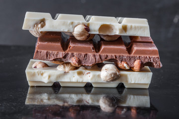 Pieces of milk chocolate with almonds and tiles of white chocolate with hazelnuts on a dark old glossy background
