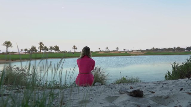 The girl is looking at game on golf course at dusk, makes selfie photo. Buggy rides through the field. Woman sits close to pond under the moon, looking at camera screen, shoots video