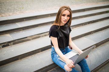 Portrait of a happy young woman sitting on the city stairs and using laptop computer