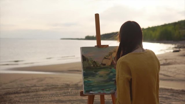 A young artist draws a palette knife still life, a girl creates a work of art near the sea or river. Person placed Malbert in the open air in order to draw from nature