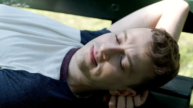 Young man lying on the bench in the park and smiling to the camera, steadycam shot
