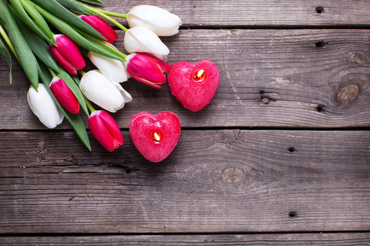 Two red  burning candles in form of  heart  and bright  spring  tulips flowers on rustic wooden background.
