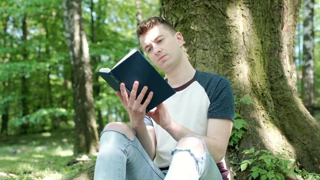 Young man looks absorbed while reading book in the forest, seteadycam shot
