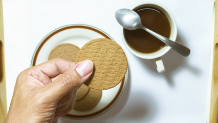 Hand hold brown biscuits in dish pairing with black coffee with teaspoon