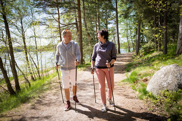 Summer sport in Finland - nordic walking. Man and mature woman hiking in green sunny forest. Active people outdoors. Scenic peaceful Finnish summer landscape. - obrazy, fototapety, plakaty
