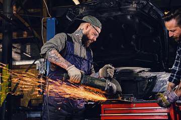 Two b mechanics working with an angle grinder in a garage. - obrazy, fototapety, plakaty
