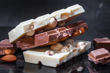 Pieces of milk chocolate with almonds and tiles of white chocolate with hazelnuts on a dark old glossy background