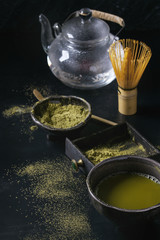 Green tea matcha powder and hot drink in black bowls and wood box standing with glass teapot, bamboo traditional tools spoon, whisk in terracotta tray over dark metal background. Close up