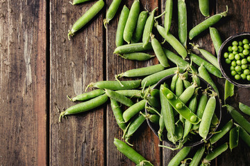 Young organic green pea pods and peas in bowls over old dark wooden planks background. Top view with space. Harvest, healthy eating.
