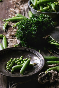 Young organic green pea pods and peas in vintage plate and bundle of parsley over old dark wooden planks with sackcloth textile background. Dark rustic style. Harvest, healthy eating.