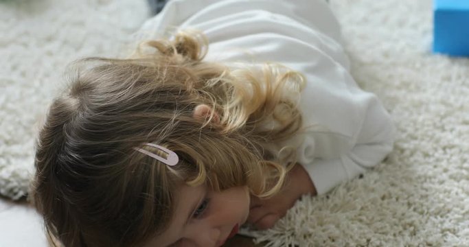 Portrait of little girl with blond curly hair lying on floor at home at the day time. Indoor.