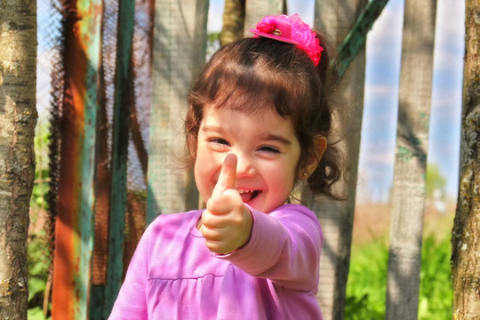 Cute smiling little girl in purple dress keeps thumbs up in warm sunny day in outdoor conditions