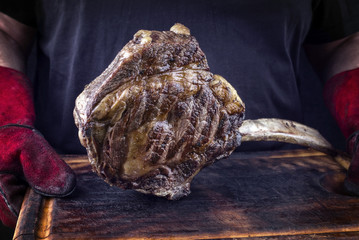 Man is holding Barbecue Wagyu Tomahawk Steak on old burnt cutting board in his hands with gloves