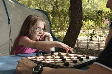 Little girl playing with her father draughts board outdoor (camping) in the sunny summer day.