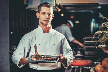 Food concept. Young handsome chef in white uniform hold in his hands ready beef steak meat ribs dish with herbs in interior of modern professional restaurant kitchen. Ready to serve. Ready to eat