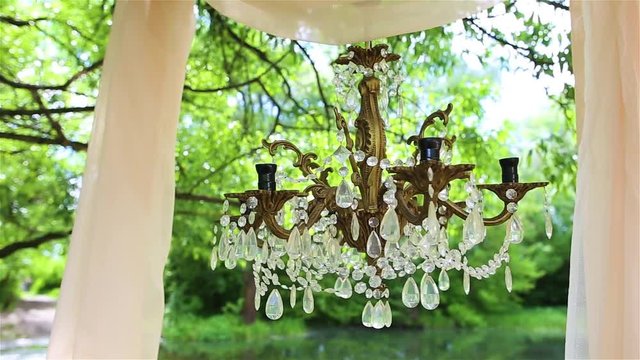 Beautiful luxury decor for outside wedding celebration. Closeup of christal chandelier without bulbs hanging in green summer park or wood. Arch decorated with draped fabric.