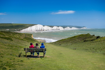 Three people sitting on a bench watching the waves on a windy but sunny day at Cuckmere Haven Sussex UK - obrazy, fototapety, plakaty
