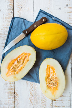 Ripe organic yellow melons, halved and whole on white plank wood garden table, pulp and seeds, blue linen napkin, knife, top view