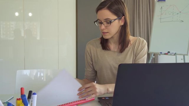 Prettty brunette girl opening journal at the table. Attractive young woman in glasses comparing information on laptop and in notebook. Cute female office worker doing her job at the desk