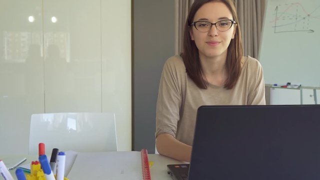 Cute caucasian girl typing on laptop at the office. Pretty young woman in glasses stopping to work on computer. Attractive brunette female manager in glasses smiling for the camera