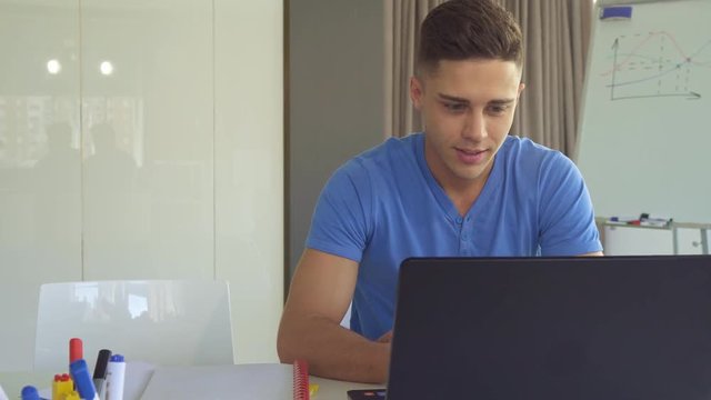 Handsome brunette guy working on laptop at the office. Attractive caucasian male manager browsing internet on computer. Cute young man in blue t-shirt looking at the screen