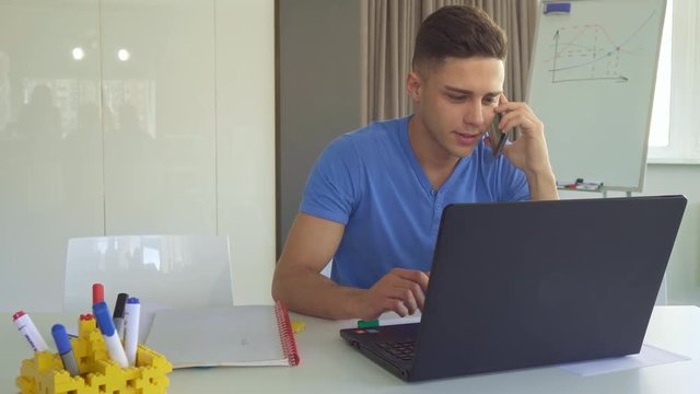 Cute caucasian guy talking on the phone at the office. Young brunette man taking his smartphone from the desk. Handsome male manager in blue t-shirt holding cellphone near his ear