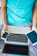 Unrecognizable man in casual clothing listening to music on smartphone and holding tablet, while reading article on laptop