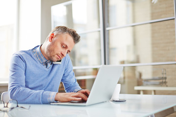 Serious middle-aged businessman typing on laptop while calling colleague on the phone, he sitting at desk in spacious office