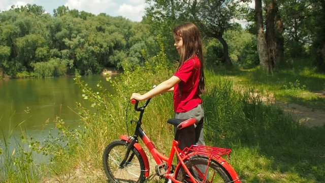 A child with a bicycle looks at the river. Little girl with a red bicycle in nature.