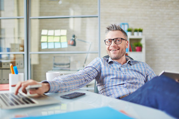 Cheerful middle-aged manager in eyeglasses working at desk and looking at colleague, he sitting at table in office