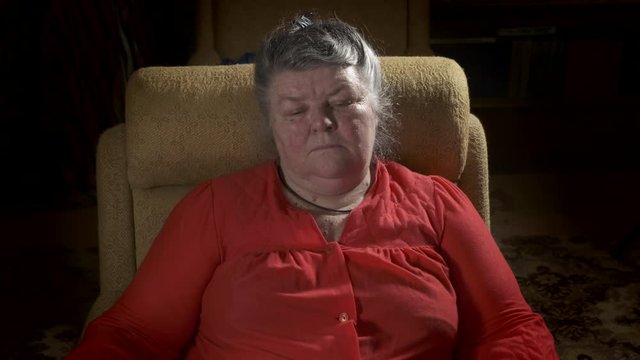 An elderly fat woman sits in armchair watching TV in a dark room at home