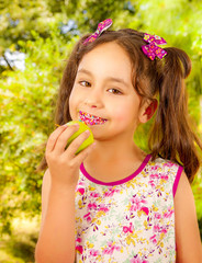 Beautiful young girl, eating a healthy apple and colorful dragees in her lips, in a blurred garden background