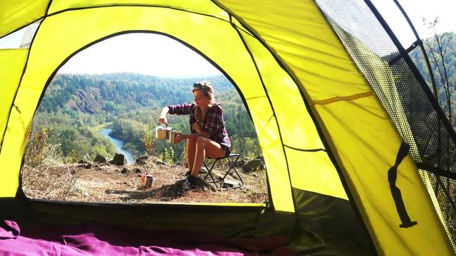 beautiful young woman sitting with metal cup on chair in camp with green forest on background 
