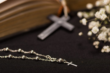Close up of a silver rosary beads and a blurred white small flowers with rosary beads over a holy bible, black background