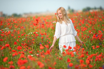 Fototapeta na wymiar blond woman in white dress walking thru blooming summer red flowers field