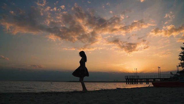 Silhouette of a girl in a light dress on the beach at sunset. Beautiful young girl walking on sand barefoot. slow motion